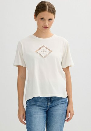 Joven con camiseta blanca de manga corta con un patrón de corte en forma de diamante, combinada con jeans azules, de pie contra un fondo liso.