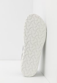 Sandale noire avec une semelle en caoutchouc texturé blanche présentant un motif en vagues, marquée du nom de la marque "Birkenstock" au bas.
