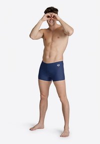 Marinblåa badshorts i stretchmaterial, med en logga på midjebandet och en figurnära design, på en manlig modell med kort hår.