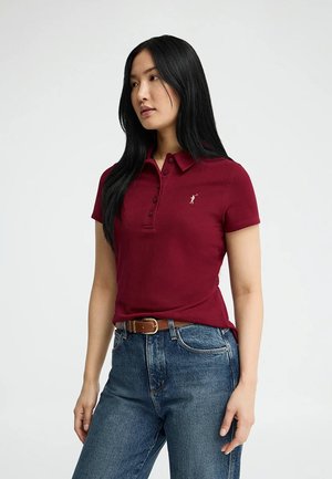 Vrouw met lang zwart haar, gekleed in een bordeaux polo en blauwe spijkerbroek met een bruine riem, licht naar links gekeerd tegen een effen achtergrond.