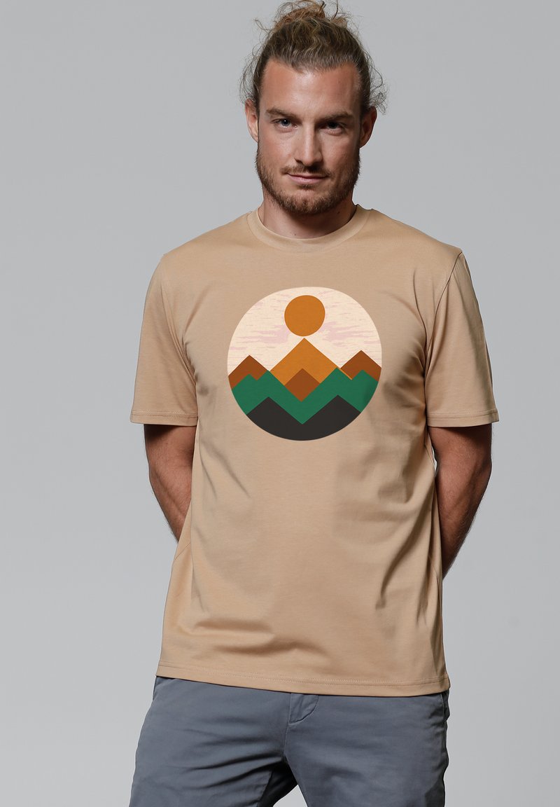 Beige T-shirt met een ronde afbeelding van bergen in bruin, groen en zwart, met een zon erboven, gemaakt van lichte stof.