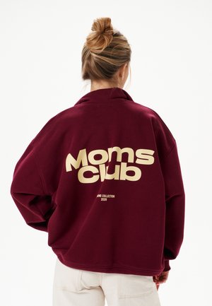 Frau mit blondem Haar im Dutt, trägt eine bordeauxrote Jacke mit der Aufschrift „Moms club“ in großen beigen Buchstaben auf dem Rücken.