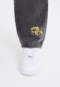 Hvide sneakers med perforeret tå og grå denim jeans, der har et guld broderet grafik af to ter på den nederste del af benet.