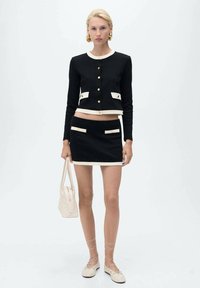 Giacca nera cropped con dettagli beige e bottoni dorati, abbinata a una mini gonna nera con due tasche frontali e bordo beige.