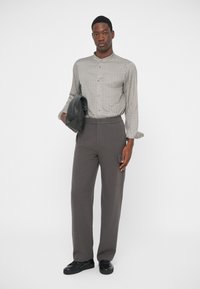 Νεαρός άνδρας με καρό μπεζ πουκάμισο και γκρι παντελόνι, κρατώντας μία μαύρη δερμάτινη τσάντα, στέκεται μπροστά σε λευκό φόντο.