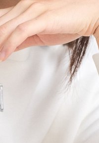 Mano levantada cerca de la cara, vistiendo una prenda blanca de manga larga, con cabello oscuro parcialmente visible en el fondo.