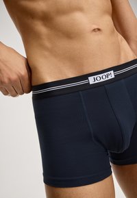 Uso ravvicinato di un uomo che indossa boxer blu navy con una fascia nera caratterizzata da strisce bianche e un patch con il logo JOOP!
