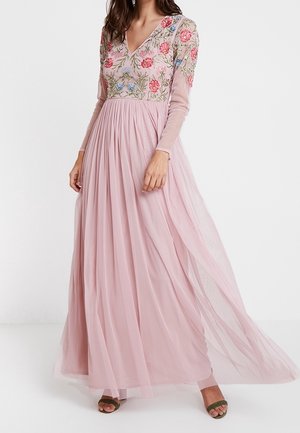 Woman wearing a long, flowing pink dress with sheer sleeves and floral embroidery on the bodice, paired with green open-toe heels.
