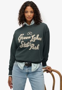 Sudadera de cuello redondo en verde oscuro con "Green Lakes State Park" y "Superdry New York" impresos en color crema, utilizada sobre una camisa de rayas clara.