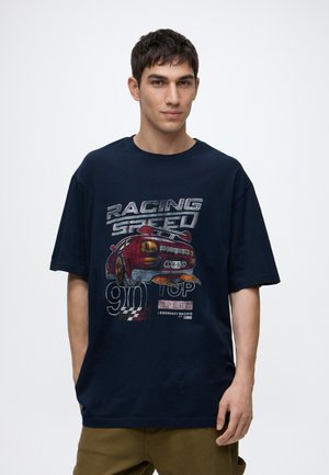 Ung mand med kort mørkt hår iført en mørkeblå T-shirt med en rød racerbils og "RACING SPEED" tekst foran.