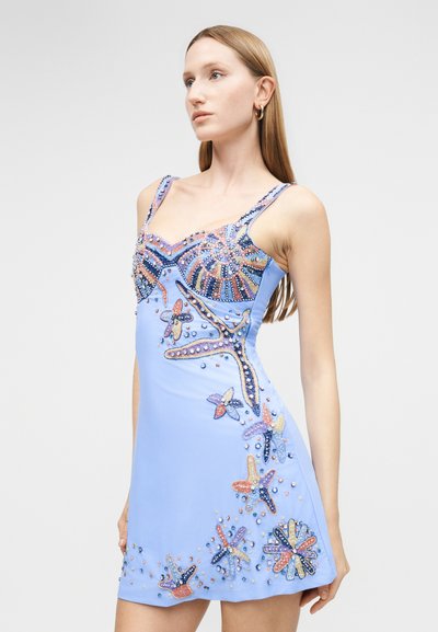 Νεαρή γυναίκα με μακριά μαλλιά που φορά αμάνικο ανοιχτό μπλε φόρεμα διακοσμημένο με πολύχρωμα, χαντρωτά σχέδια αστερία και κοχυλιών.