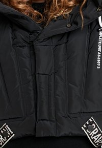 Svart vadderad jacka med hög krage, med kviltad textur, tryckknappar och en dekorativ märke med mönstrad kant.