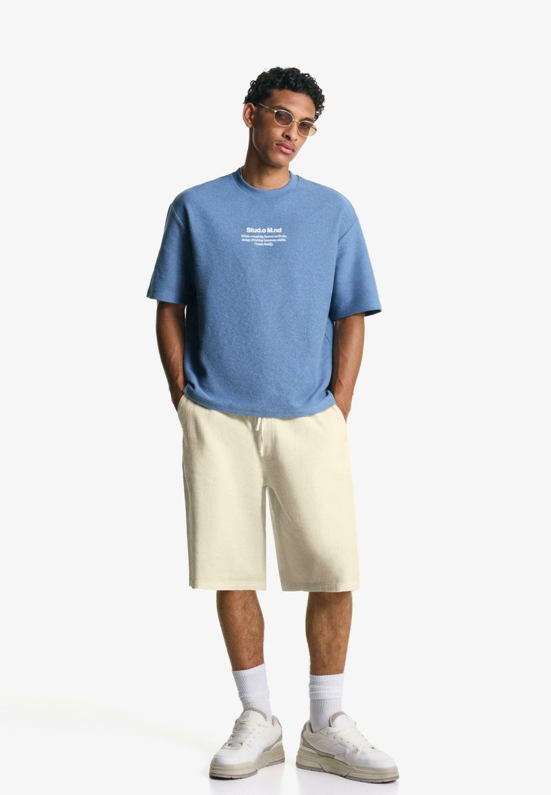 T-shirt blu in cotone con maniche corte e testo bianco. Pantaloni corti chiari in tessuto morbido, abbinati a sneakers bianche e calzini alla caviglia.