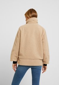 Beige fleecejakke med høj krave og oversized pasform. Har lange ærmer og en struktureret, blød overflade.