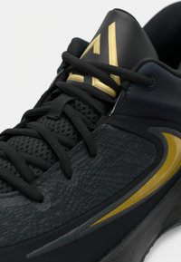 Sort og guld atletiksko med struktureret stof, sorte snørebånd og et guld swoosh-logo på siden, vist i nærbillede.