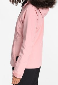 Person, der eine hellrosa Winterjacke mit einem gefütterten Kapuze trägt, die zugezippt ist, und schwarze Hosen, seitlich gezeigt.