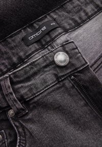 Gros plan sur un jean en denim noir montrant un bouton métallique gravé de "OMBRE DENIM" et une étiquette en tissu noire avec "ombre" et la taille M/32.