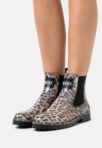 Botines tobilleros con estampado de leopardo hechos de goma transparente, con paneles laterales elásticos negros y un logo de la marca en el frente. Diseño con suela plana.
