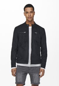 Svart semsket skinnjakke med ståkrage, glidelås i brystlommene og en slank, tettsittende design. Båret over en hvit strikket skjorte og grå shorts.