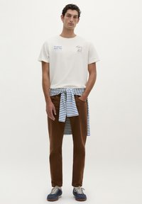 Weißes Baumwoll-T-Shirt mit blauen Grafiken, braune Hose und ein blau-weiß gestreiftes Hemd, das um die Taille gebunden ist. Sneaker mit braunen Sohlen.