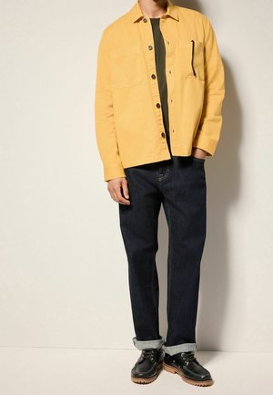 Gul bomuldsskjortejakke over en grøn t-shirt, kombineret med mørke denimjeans og sorte sko med brune såler.