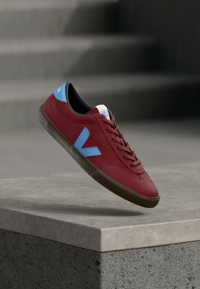 Baskets rouges avec un logo "V" bleu et un détail au talon, semelle marron, suspendues au-dessus d'un rebord en béton gris avec des escaliers en arrière-plan.