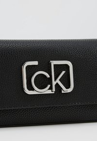 Czarny portfel z teksturowanej skóry z metalicznym logo "CK" w kolorze srebrnym. Posiada smukły, prostokątny kształt oraz wykończenie ze stębnowania na krawędziach.