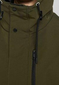 Detail pohledu na olivově zelenou bundu s černým zipem, patentními knoflíky a černými stahovacími šňůrkami na kapuci.
