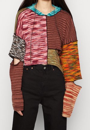 Femme portant un pull court en patchwork avec des manches longues dépareillées et un pantalon noir taille haute sur un fond uni.