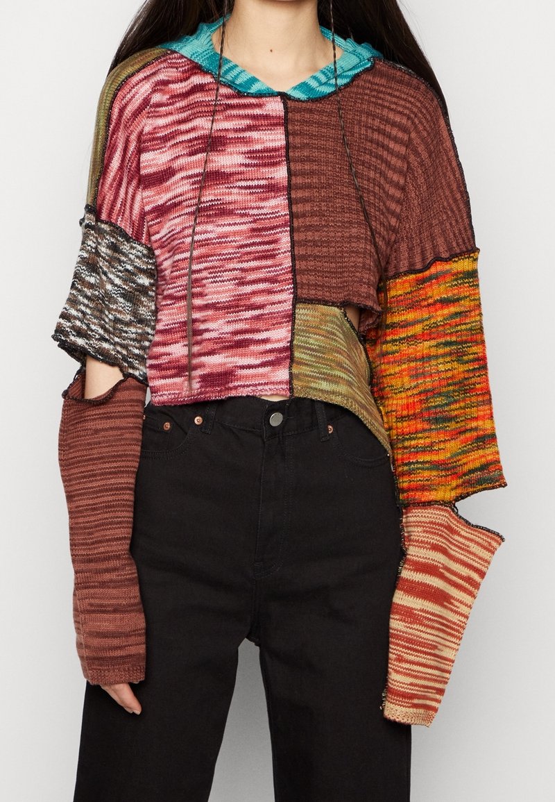 Femme portant un pull court en patchwork avec des manches longues dépareillées et un pantalon noir taille haute sur un fond uni.