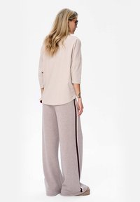 Vrouw met lang blond haar draagt een beige losse top, grijze wijde broek met een donkere zijstreep, zonnebril en oorbellen met een ring, staande in profiel.