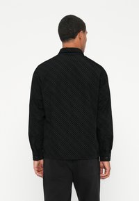 Juodas ilgomis rankovėmis marškinėliai su tekstūruotu raštu, turintys apykaklę ir mygtukais užsegamas rankoves. Audinys atrodo minkštas ir šiek tiek pakeltas.