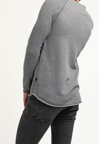 Maglietta grigia a maniche lunghe con una trama a maglia testurizzata, orlo arrotondato e dettagli sfrangiati; indossata con jeans neri, che mettono in mostra un design casual.