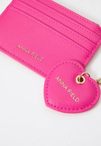 Pink synthetic card holder with multiple slots and a heart-shaped keychain, both featuring gold lettering "ANNA FIELD." Textured finish.