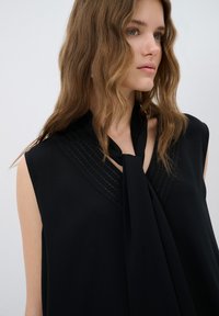 Haut sans manches noir avec un lien au niveau du col, présentant un motif texturé subtil et un tissu délicatement drapé. Matériau lisse et léger.