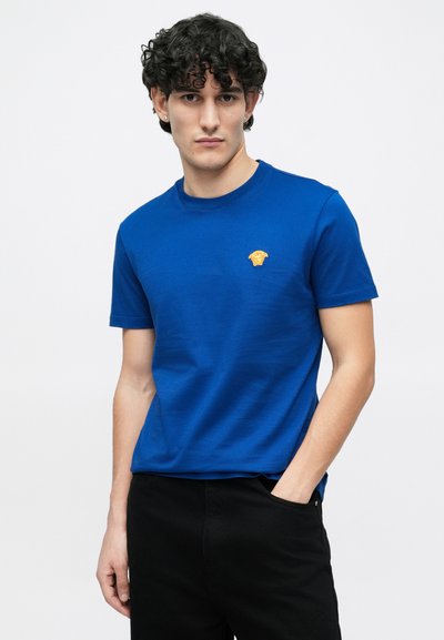 Kék pamut anyagú kerek nyakú póló, melyen a mellrészen egy apró arany embléma található. Rövid ujjú, klasszikus szabású, fekete nadrággal párosítva.