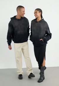 Deux individus portent des sweats à capuche assortis dans un dégradé de noir à gris foncé, avec des poches kangourou. L'un porte des bottes noires à chevilles et l'autre des pantalons crème.