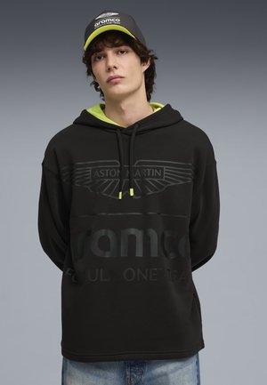 Junger Mann, der einen schwarzen Hoodie und eine passende Kappe des Aston Martin Aramco Formel-1-Teams vor einem schlichten grauen Hintergrund trägt.