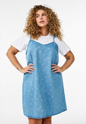 Denim dress in light blue with a floral pattern, adjustable shoulder straps, and a relaxed fit. Worn over a white T-shirt.