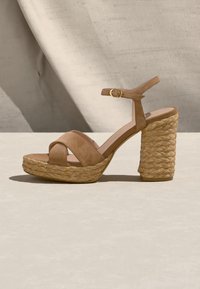 Sandale à bout ouvert en daim couleur cognac avec semelle plateforme tissée, talon bloc et bride de cheville avec boucle dorée sur fond neutre.