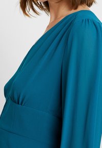 Türkises Kleid mit tiefem V-Ausschnitt und gerafften langen Ärmeln. Der Stoff wirkt weich mit einer glatten Textur und hat eine taillierte Passform.