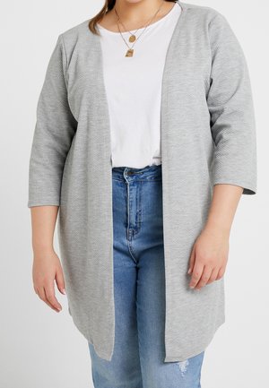 Vrouw draagt een lichtgrijze openvallende cardigan, wit shirt, blauwe spijkerbroek en gelaagde gouden kettingen tegen een effen achtergrond.