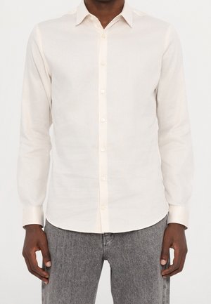 Mand iført en hvid skjorte med lange ærmer og knapper og grå jeans, stående foran en ensfarvet lys baggrund.
