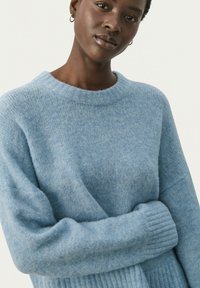 Pull en maille bleu clair avec un col rond, coupe décontractée et poignets côtelés. Texture douce, finition légèrement duveteuse, sans motifs ni accents visibles.