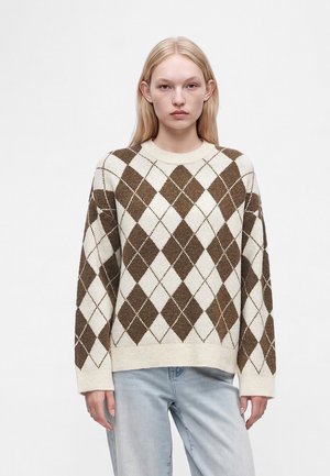 Jeune femme aux cheveux blonds raides porte un pull à motif losanges crème et marron avec un jean bleu clair sur un fond blanc uni.