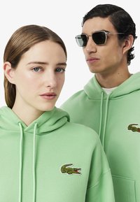 Due persone con felpe pastello verdi abbinate, complete di cordini. Ognuna ha un logo di coccodrillo ricamato verde sul petto.