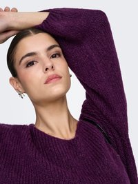 Maglione viola lavorato a maglia con superficie strutturata, scollo rotondo e spalle scese, abbinato a orecchini d'argento e una collana semplice.