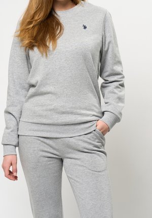 Kvinde iført lysegrå sweatshirt og matchende sweatpants med venstre hånd i lommen mod hvid baggrund.