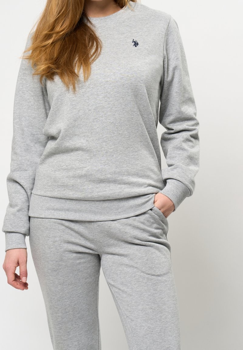 Femme portant un sweat-shirt gris clair et un pantalon de survêtement assorti, avec la main gauche dans la poche, sur fond blanc.