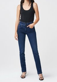 Jeans de mezclilla azul con un corte ajustado, cintura alta, diseño clásico de cinco bolsillos y lavado oscuro. Combinados con una camiseta sin mangas negra y sandalias.